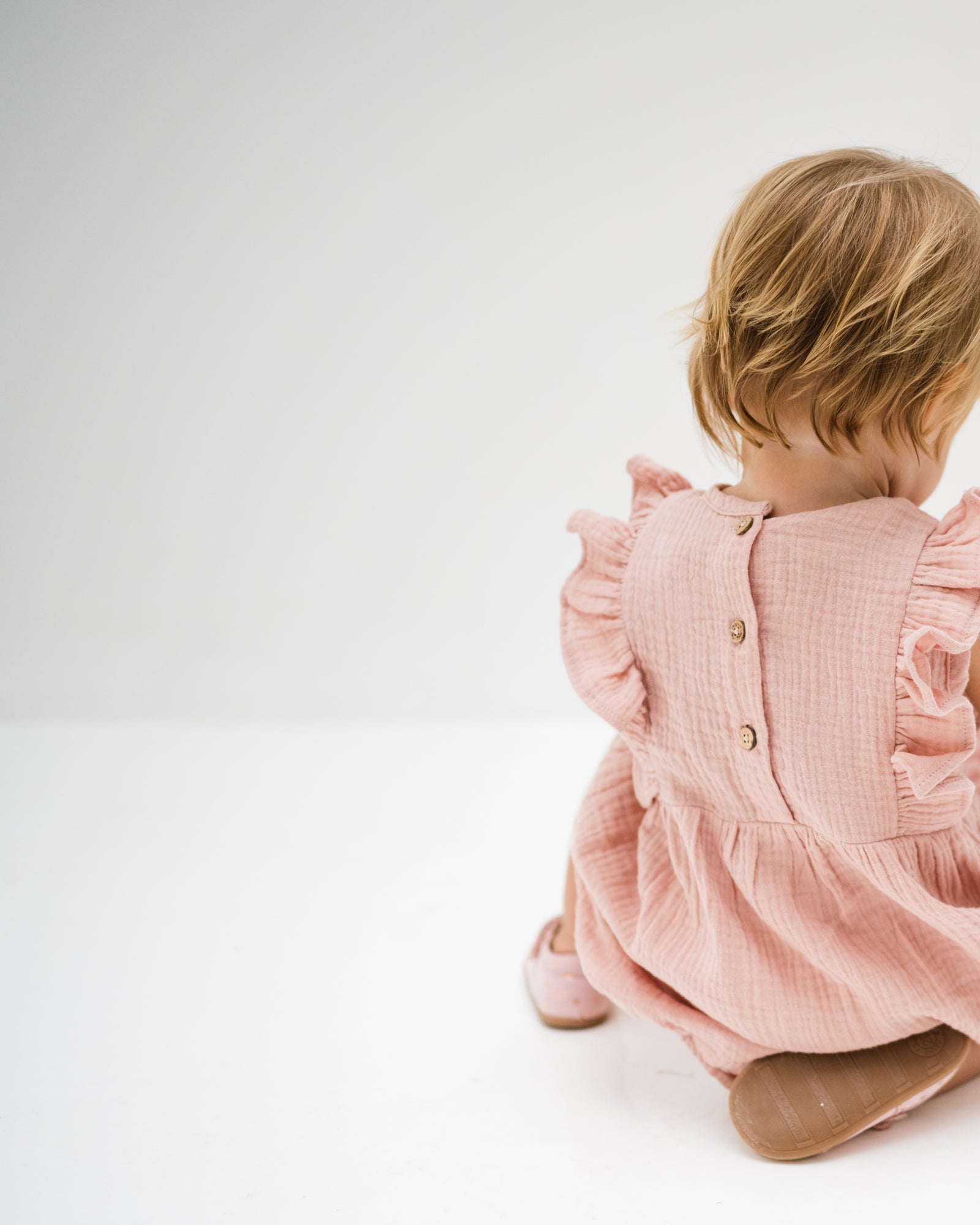 baby girl of 12 months showing the frilled Rose romper in soft pink muslin from the back