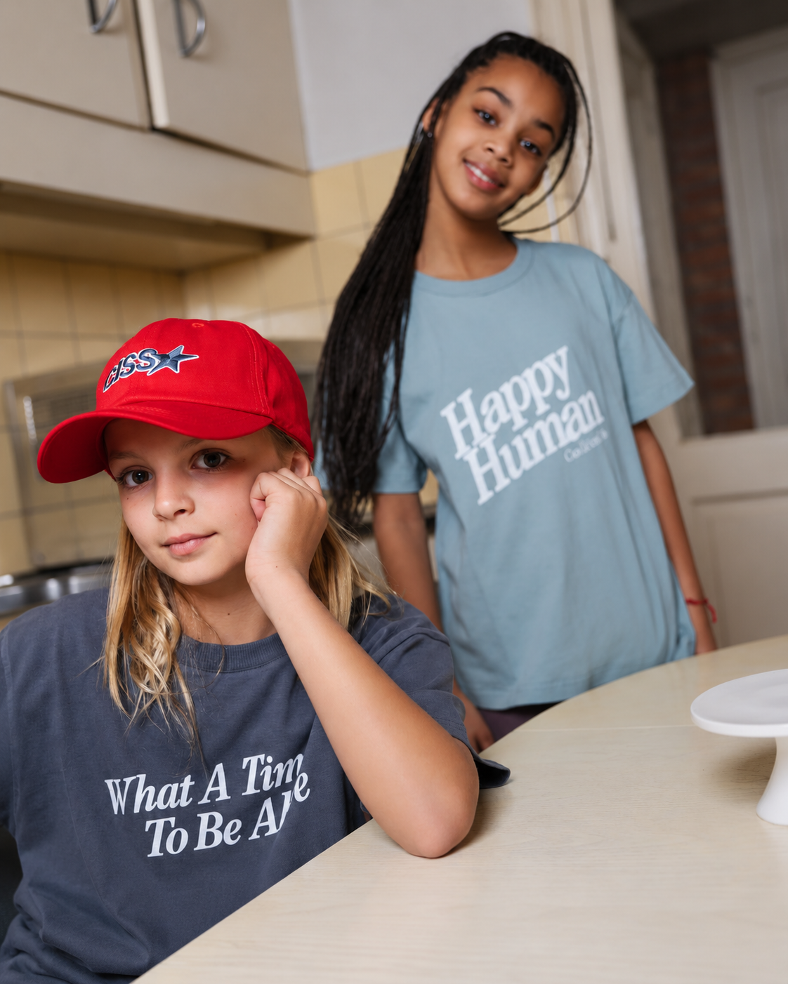 Blond teenage girl wearing the What a time to be alive t-shirt in turbulence in organic cotton  and the other girl wearing Happy Human t-shirt in silver blue;