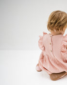 baby girl of 12 months showing the frilled Rose romper in soft pink muslin from the  back