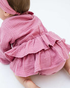 baby girl wearing the Helena onesie in blush pink muslin with long sleeves, showing the back
