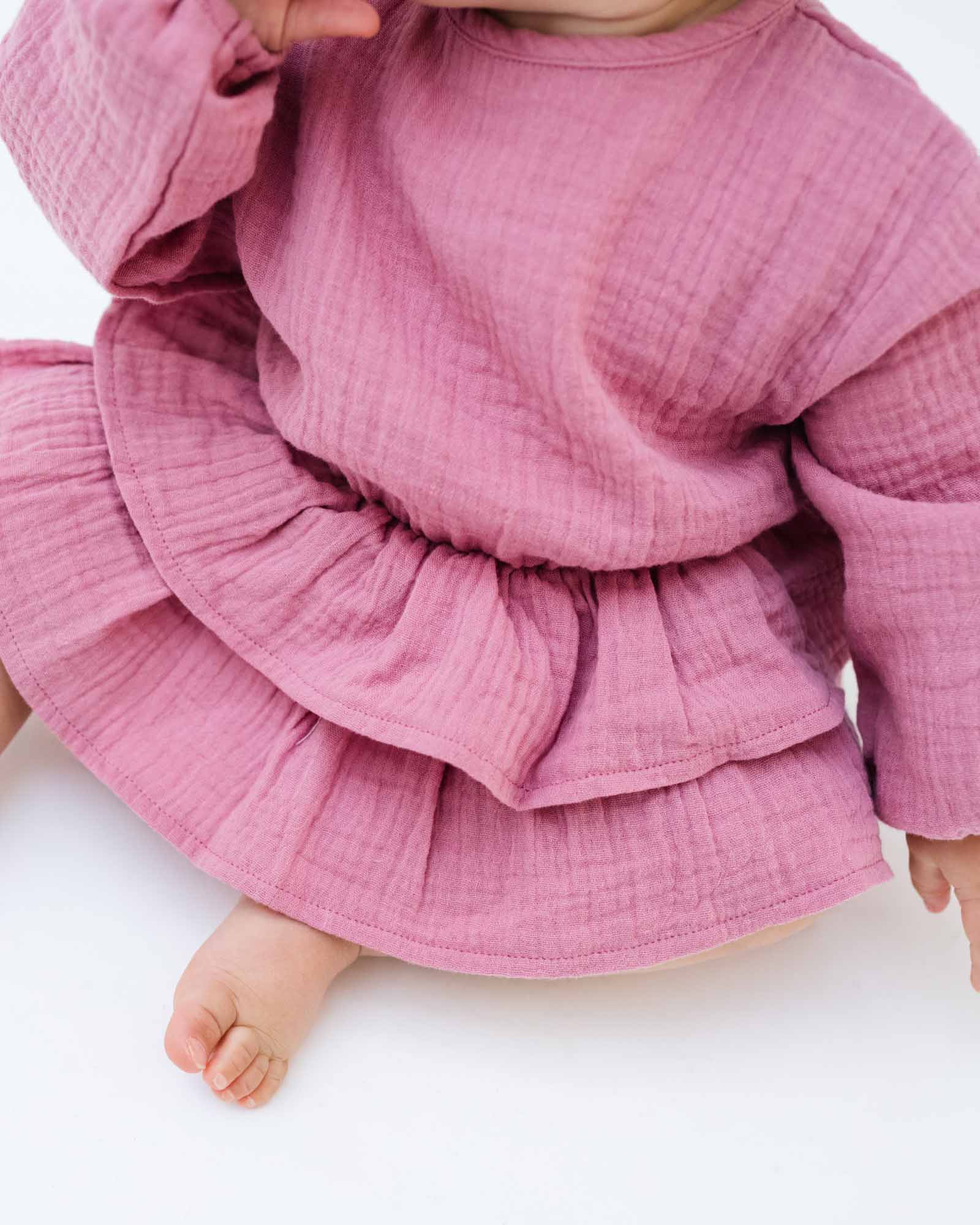 baby girl wearing the Helena onesie with long sleeves in blush pink muslin cotton, showing the 2 layers of frills around the waist