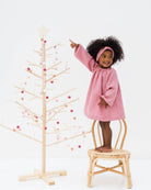 smiling 2-year-old girl looking at the christmas tree, wearing the romane dress with long sleeves in rose muslin cotton with golden stars together with the matching twisted headband