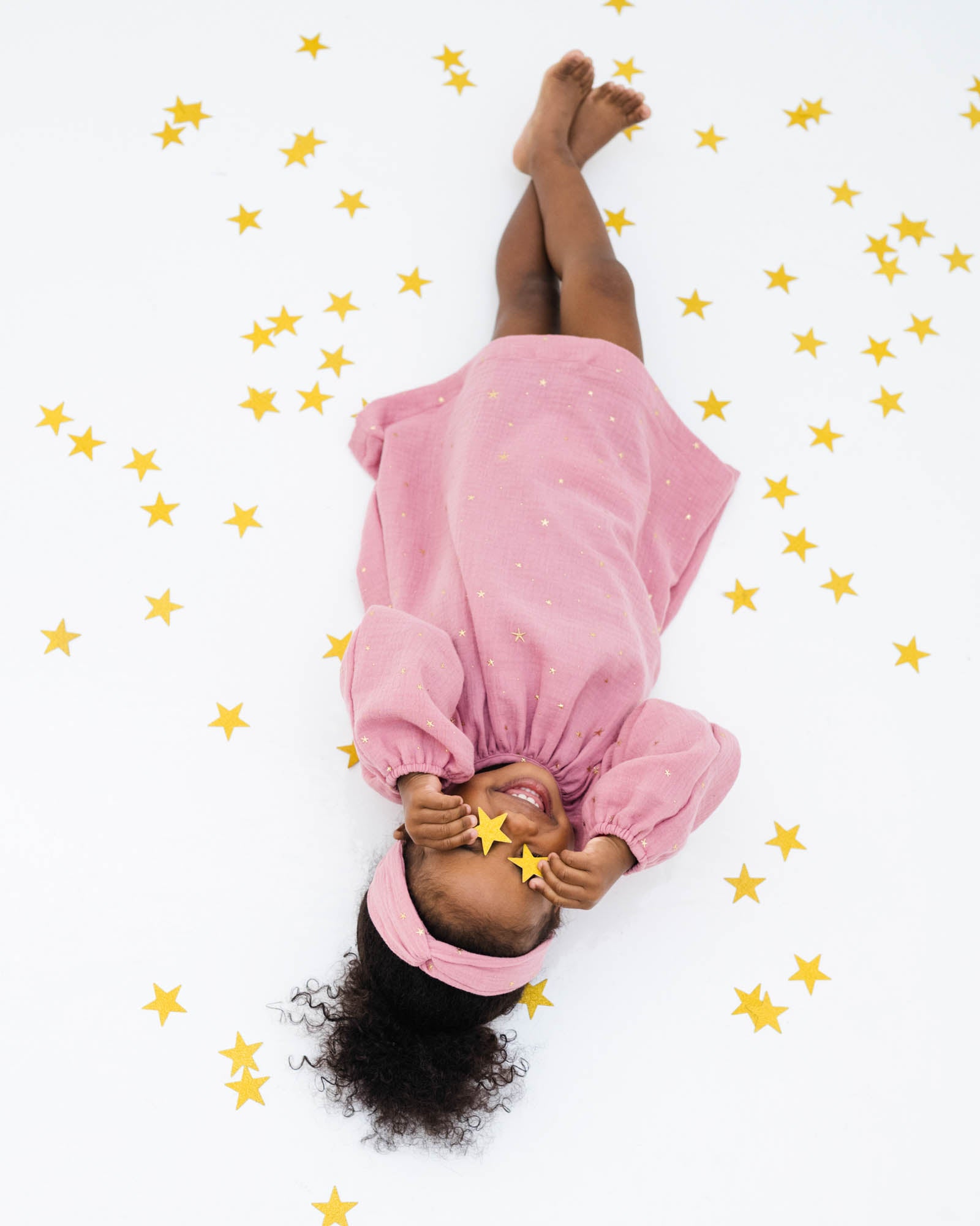 sassy 2-year-old girl laying on the floor between stars, wearing the romane dress with long sleeves in rose muslin cotton with golden stars