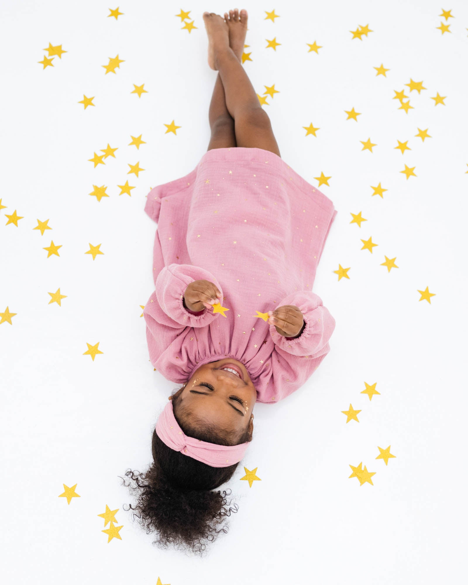 2-year-old girl laying on the floor between stars, wearing the comfortable and stylish romane dress with long sleeves in rose muslin cotton with golden stars