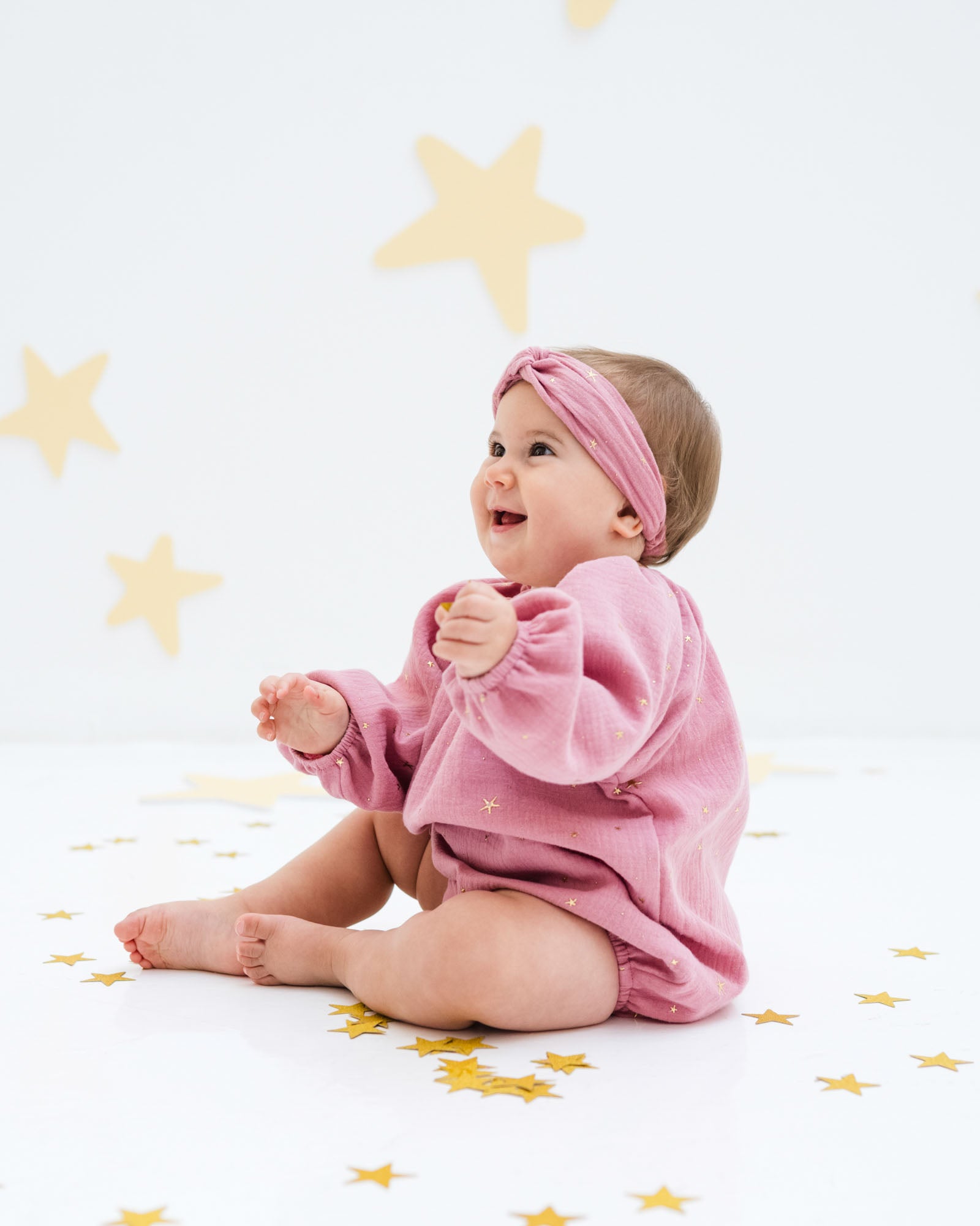 baby girl is wearing the Romane onesie with the matching twisted headband in rose pink with golden stars