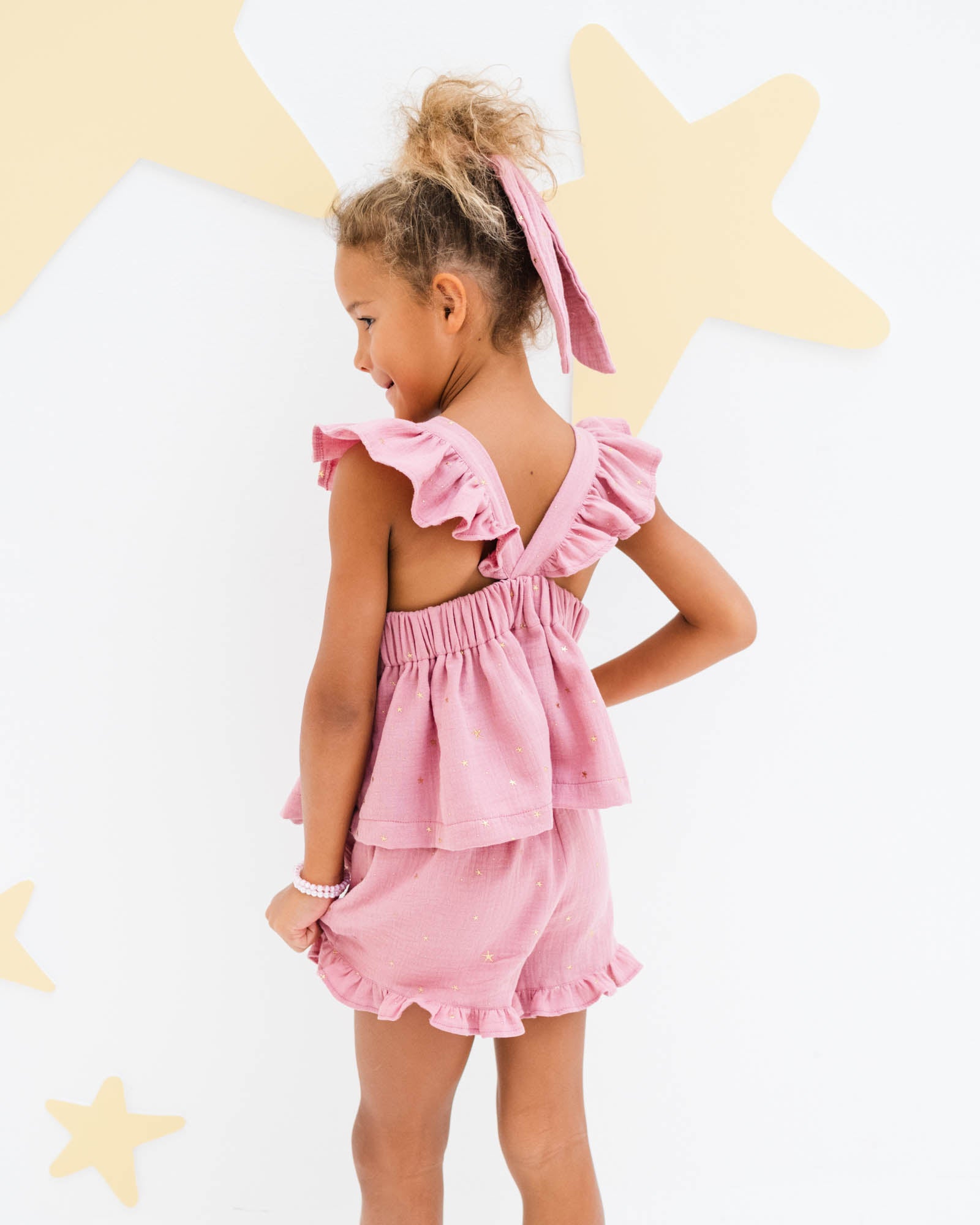 young girl showing the back of the frilled sleeveless Rosalie top and Charlene frilled  shorts in rose pink muslin cotton with golden stars 