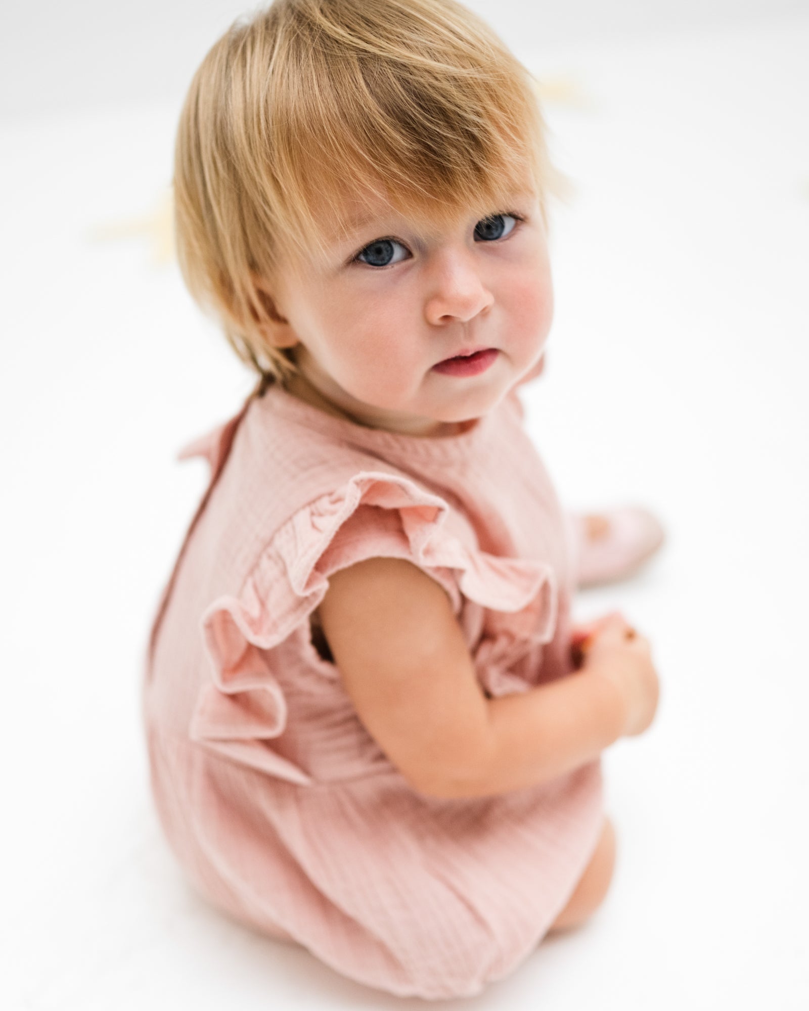 baby girl of 12 months showing the frilled Rose romper in soft pink muslin from the side 